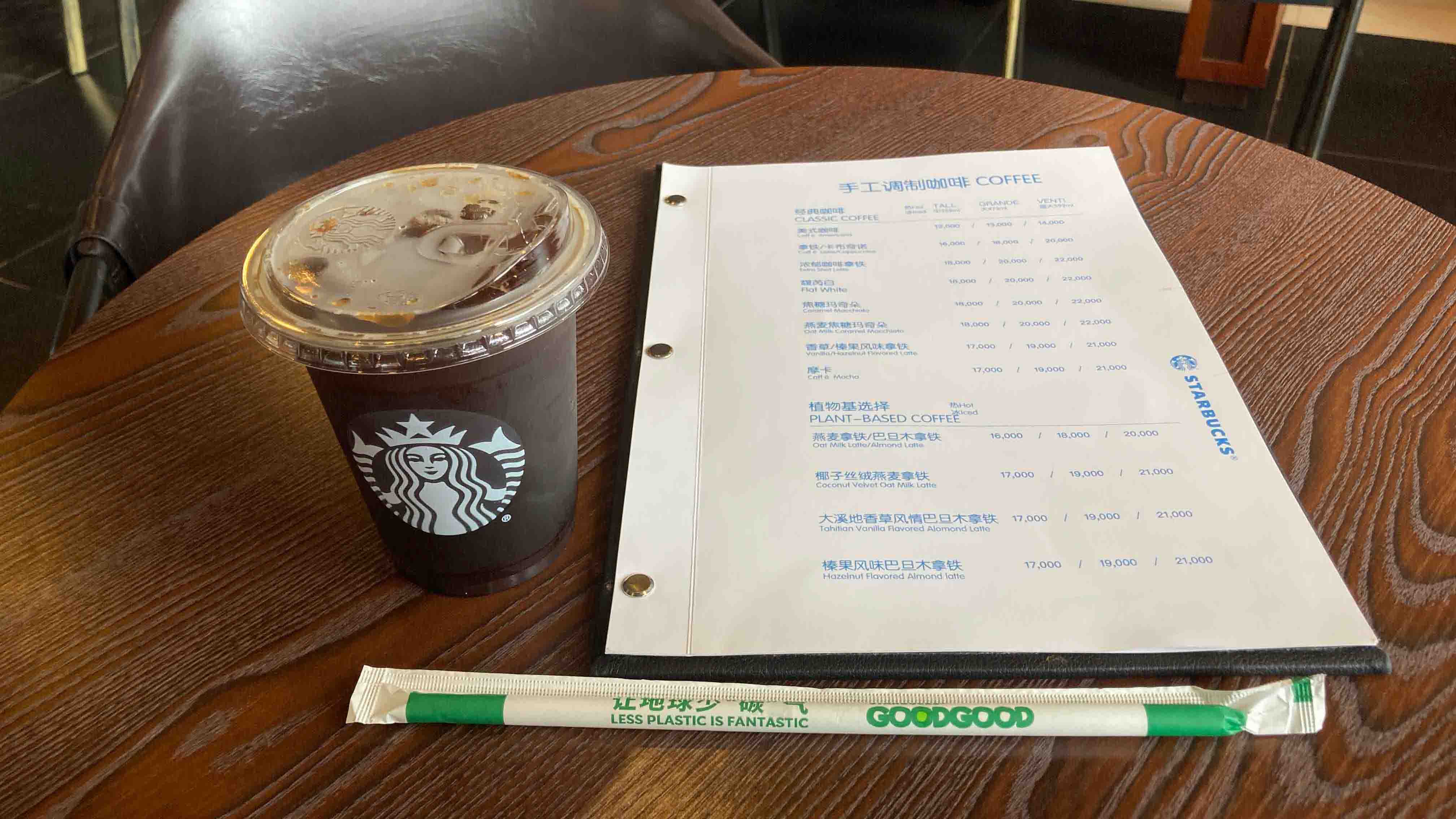 Starbucks iced coffee on a wooden table next to a bilingual menu and an eco-friendly straw labeled “LESS PLASTIC IS FANTASTIC.”