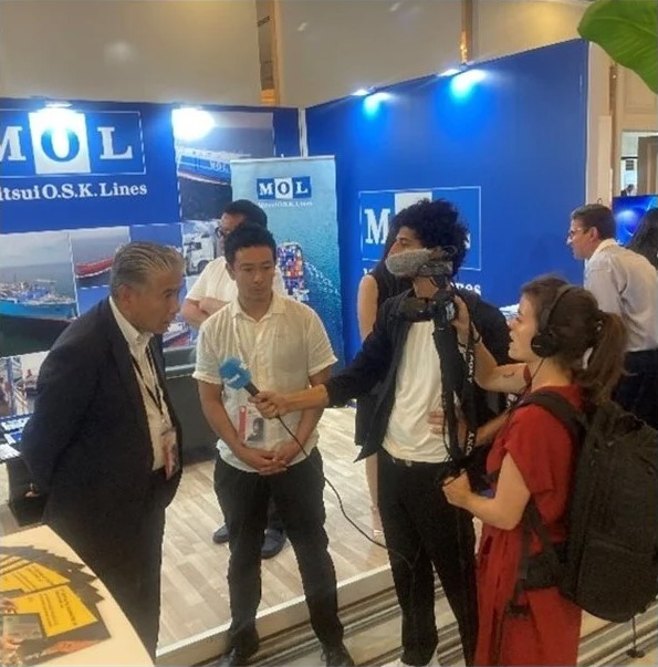 展示会ブースでインタビューを受けるMOL（商船三井）のスタッフ。背景にはMOLのロゴと展示パネルが見える。