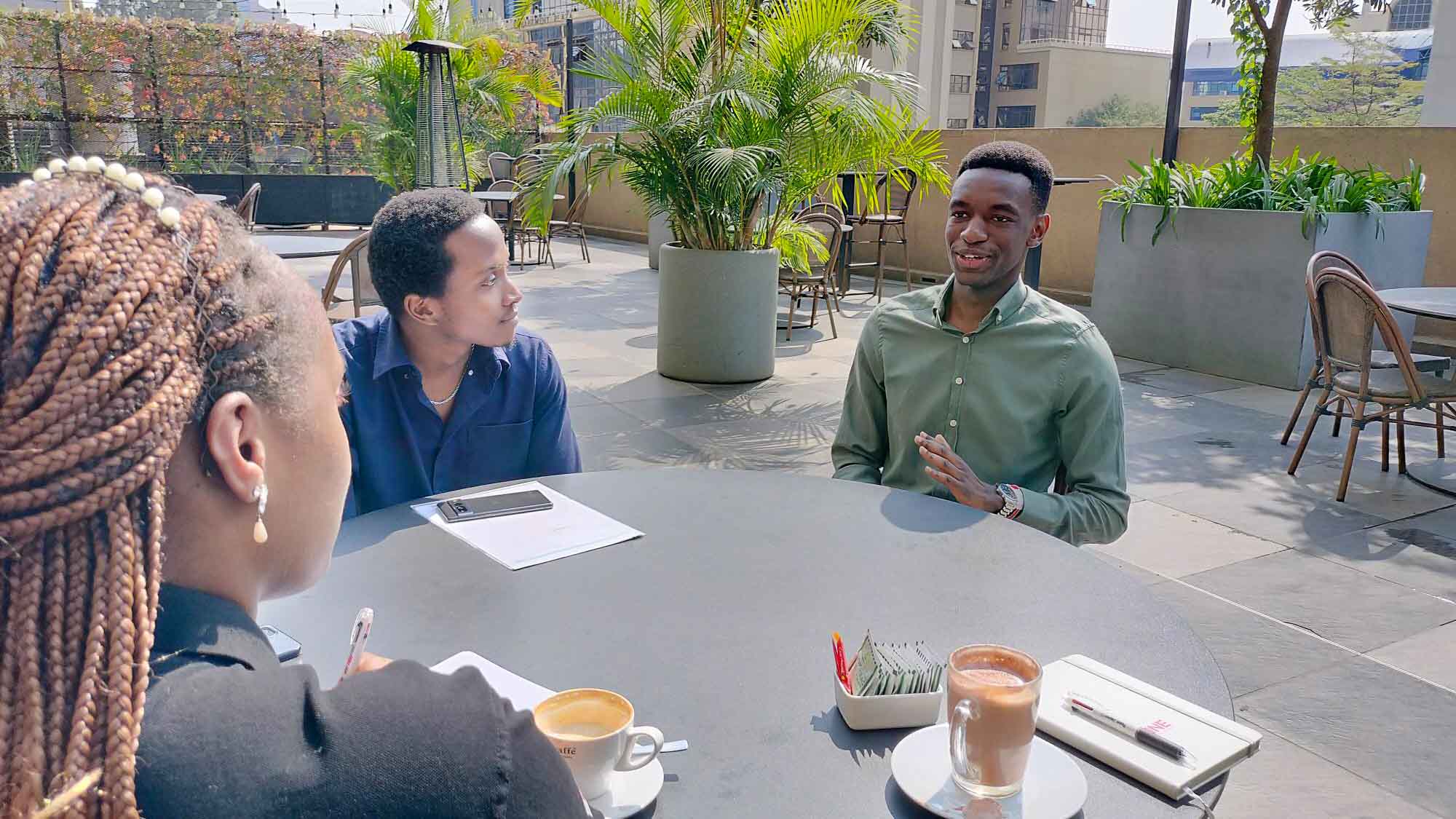日本語学習の体験について語る3人のケニア人学習者。屋外のカフェテーブルで、ノートやコーヒーを前に話し合っている様子。