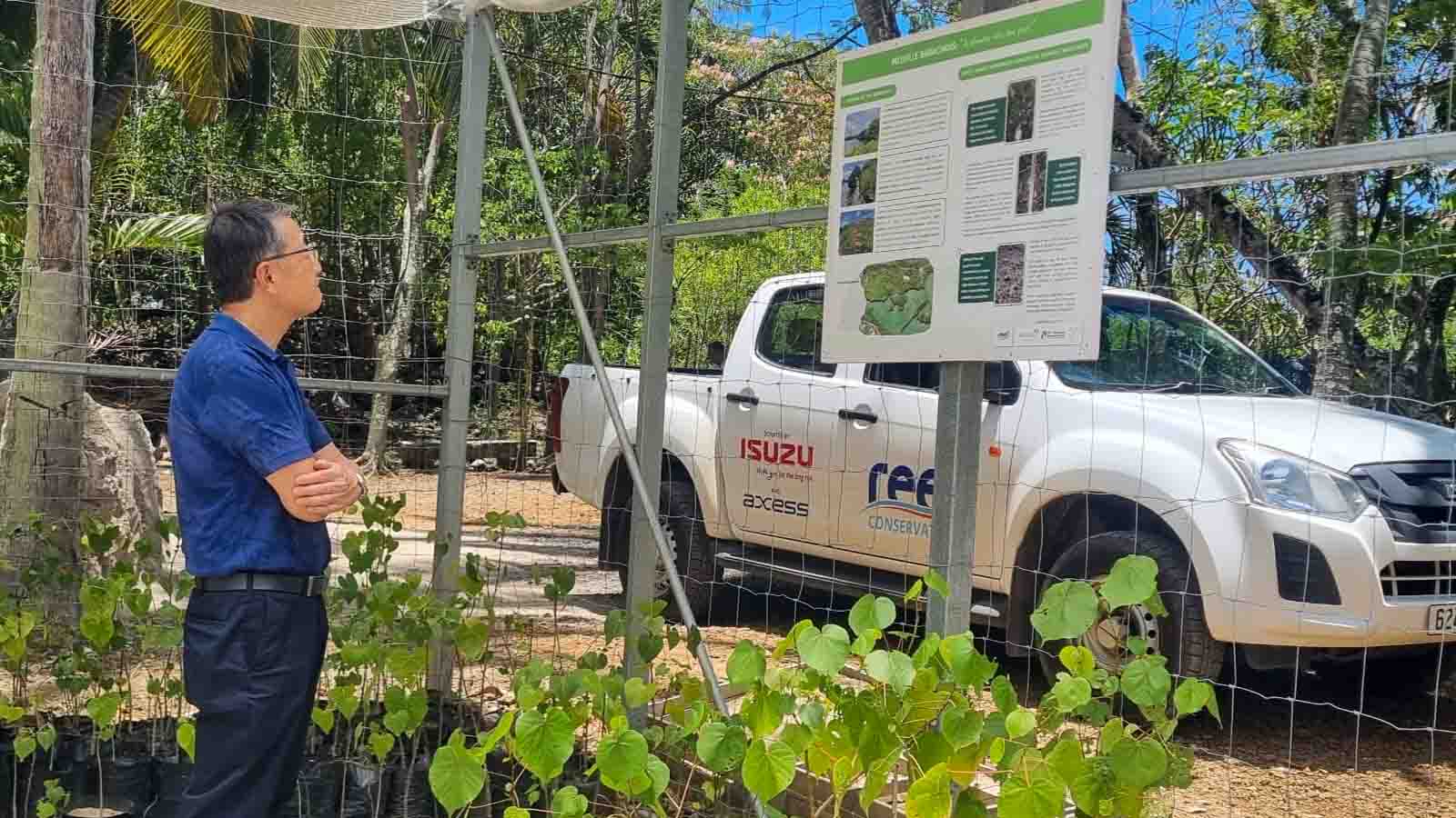 MOL Chairman Junichiro Ikeda, observing an informational display at a nursery site in Mauritius, with young plants growing in the foreground and a conservation vehicle in the background.